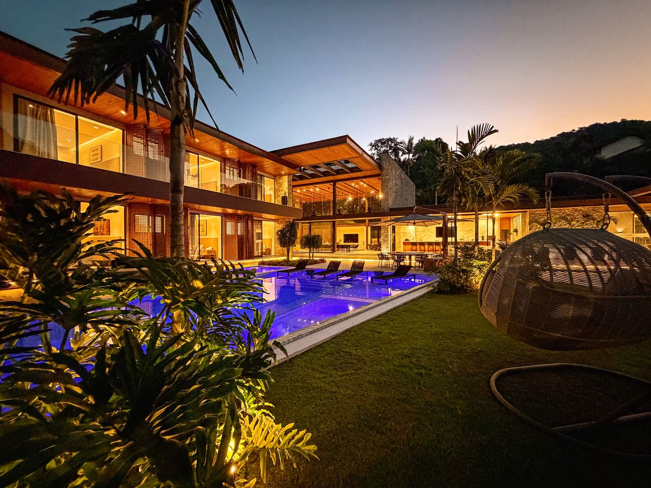 A residência iluminada ao anoitecer: fachada em muxarabi acesa em camadas de luz, refletida na piscina