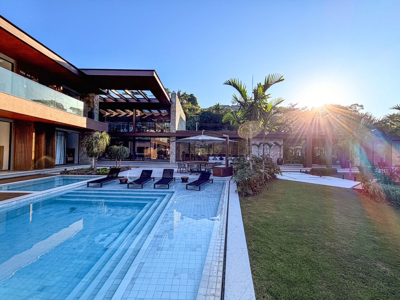 Piscina de grandes proporções com gramado e pergolado sob o sol da tarde